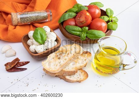 Fresh Tomatoes And Green Basil In A Wooden Bowl, Near Mozzarella, Canola Oil And Spices On A White T