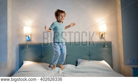 Happy Laughing And Smiling Toddler Boy In Pajamas Skipping And Jumping Up High On Parents Bed At Nig