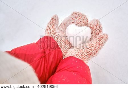 Women Hands In Red Mittens Hold A Heart Made Of Snow, Close-up. Woman Holding A Heart Shaped Snowbal