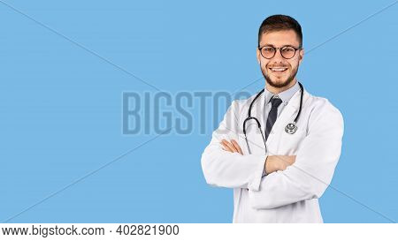 Professional Doctor Man In White Uniform Posing Crossing Hands Standing Over Blue Studio Background,