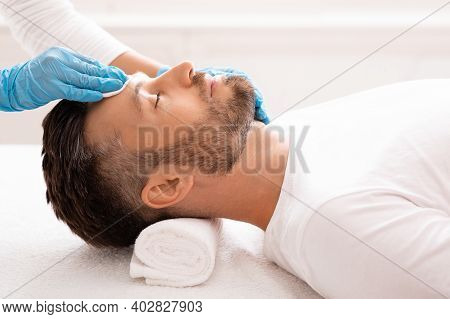 Side View Of Unrecognizable Cosmetologist Cleansing Bearded Man Face With Toner Before Or After Trea