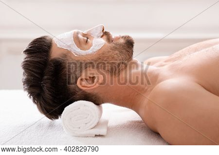 Male Cosmetology Concept. Relaxed Bearded Man With White Face Mask On Laying On Massage Table At Spa