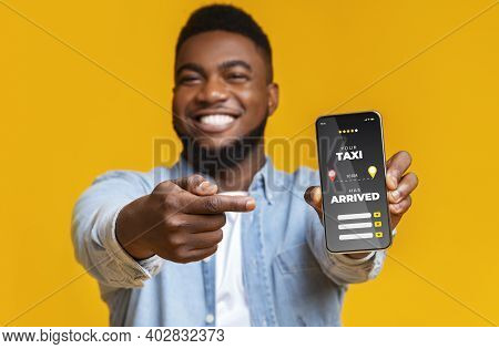 Joyful Black Guy Pointing At Smartphone With Opened App For Taxi Rate, Cheerful African Man Enjoying