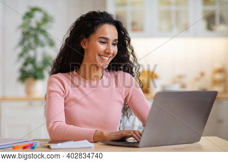 Happy Beautiful Young Female Freelancer Working On Laptop In Kitchen. Smiling Brunette Lady Typing O