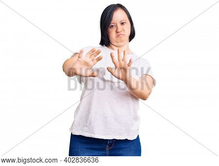 Brunette woman with down syndrome wearing casual white tshirt moving away hands palms showing refusal and denial with afraid and disgusting expression. stop and forbidden. 