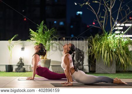 Two asian women in sportswear pants practicing yoga during city lockdown in their apratment with distance in background of cityscape illumination at night. Work out fitness healthy lifestyle concept.