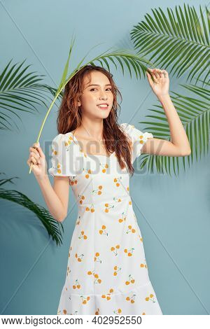 Portrait Of Stylish Serious Girl Wears Summer Dress Bahind The Tropical Leaves Over Blue Background.