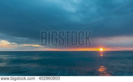 Dense Blue Clouds And Turquoise Sea. Between The Sky And The Water, The Setting Sun Illuminates The 