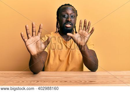 Handsome young black man wearing casual clothes sitting on the table afraid and terrified with fear expression stop gesture with hands, shouting in shock. panic concept. 