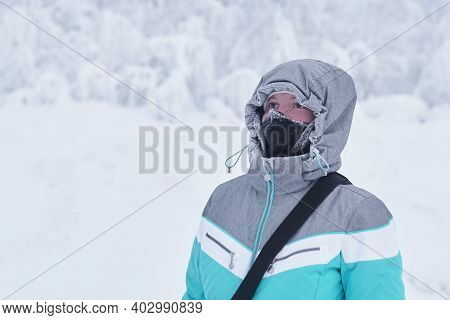 Young Woman In A Warm Jacket With A Hood And A Buff Neck Gaiter On Her Face Against The Background O