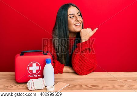 Beautiful hispanic woman with first aid kit pointing thumb up to the side smiling happy with open mouth 