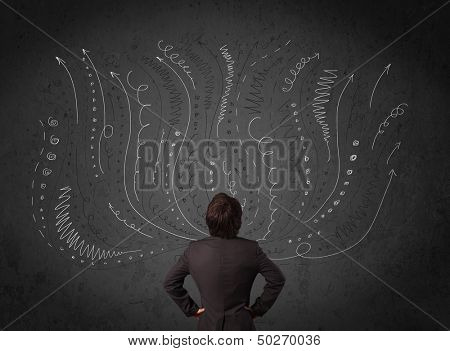 Thoughtful young businessman standing and deciding in front of a chalkboard with sketched arrows and lines in different directions
