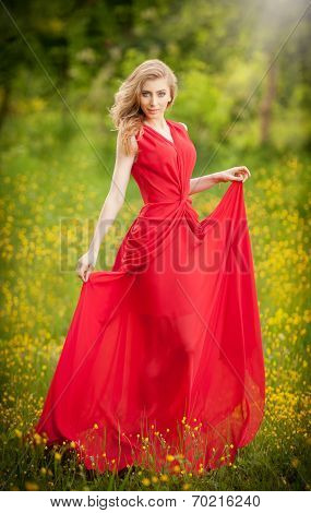 Portrait of young beautiful blonde woman wearing a long red elegant dress posing in a green meadow.