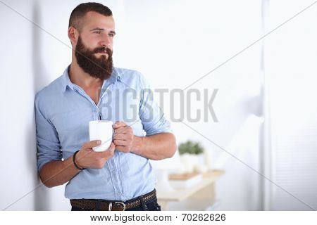 Young man  standing near wall and holding cup of coffee in offi