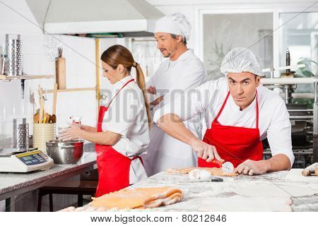 Portrait of confident male chef cutting ravioli pasta with colleagues working in background at commercial kitchen