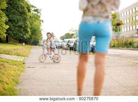 Smiling Girl Rides Her Bike On City Sidewalk As Mother Watches