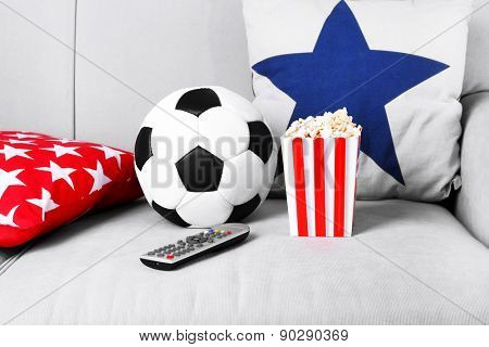 Soccer ball, remote control and box of popcorn on comfortable sofa, indoors