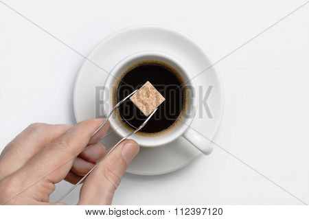 Male Hand Holding Sugar Tongs With Cane Sugar Cube Over Cup Of Black Coffee Against White Background