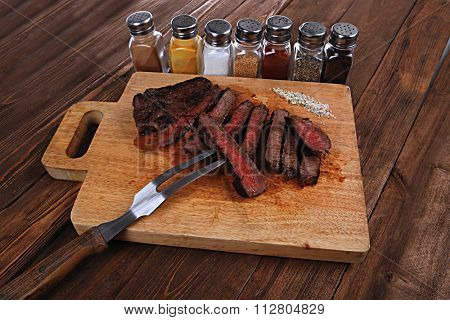 grilled beef fillet steak on fork over wooden board served with different kind of dry spices salt crushed black pepper hot baked sweet paprika red hot chili on dark table