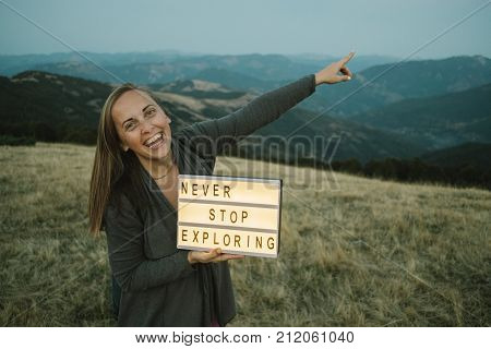 Smiling Woman With Box With Text Never Stop Exploring  Against The Background Of The Mountain. Trave