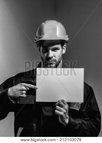 Serious man handsome builder construction mason worker repairman craftsman foreman bearded male in boilersuit and orange hard hat keeps blank paper for copy space in hands on grey background