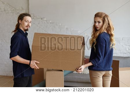 Sideways portrait of fashionable girl with long wavy hair holding large cardboard box together with her stylish boyfriend with flesh tunnels beard and dreadlocks while moving into new apartment