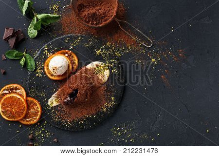 Exquisite french dessert. Chocolate fondant with creme anglaise and vanilla ice cream on round slate with orange citrons. Black background with cocoa, mint and sieve, top view, copy space