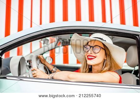 Poortrait of a young happy woman driving a car on the red wall background during the summer vacation