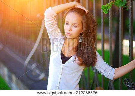 Portrait of a young pensive cute brown-haired girl standing near the rail fence in a Park and hand combing beautiful long hair on blurred background, close up.