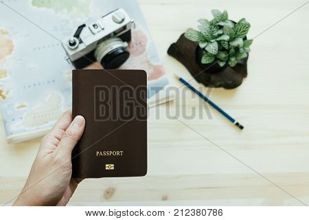 Hand Of Women Hold Passport Has Retro Camera, World Map, Pencil, Plants Placed On Wooden Table Are B