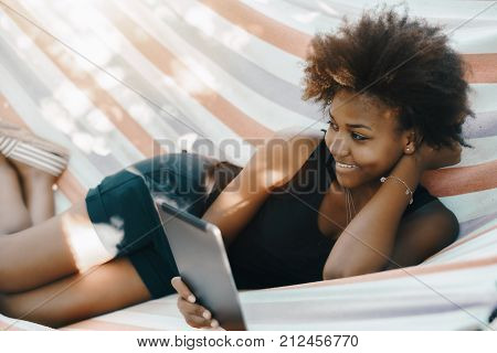 Charming young cheerful African American lady is laying on hammock outdoor in the garden smiling and looking aside while reading an internet article on her digital tablet on sunny summer evening
