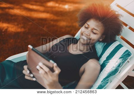 Cheerful Brazilian female laying on a deck chair and watching movie on her digital tablet; young black girl with afro laying hair on recliner and messaging via digital pad; vivid colors in background