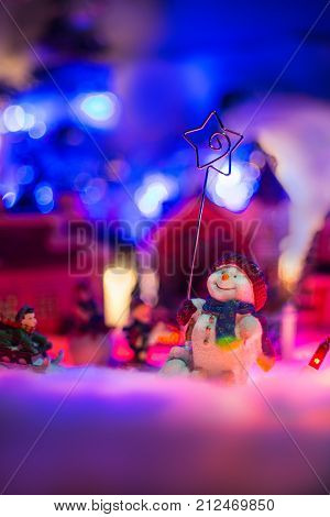 happy snowman holds star of bethlehem stands in front of colorful snowy village in the background. christmas festive miniature scenery