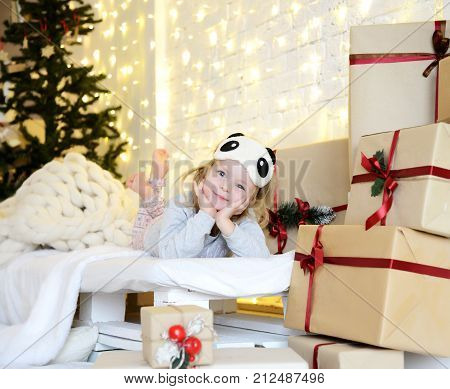 Young baby girl sitting dreaming near magical New year craft gifts by a Christmas tree on yellow magic lights background