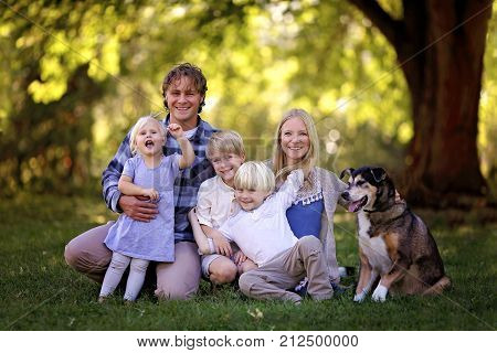 A portrait of a family of 5 blonde haired caucasian people sitting outside under the trees with their adopted pet German Shepherd Mix Breed Dog.
