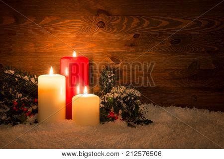 Christmas candles with Holly and conifer branches on an artificial snow and wooden background.