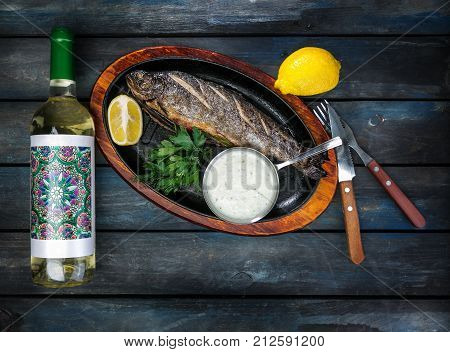 Frying pan with tasty trout fish on wooden background with a bottle of wine greens, sauce and lemon. Served with a knife and fork