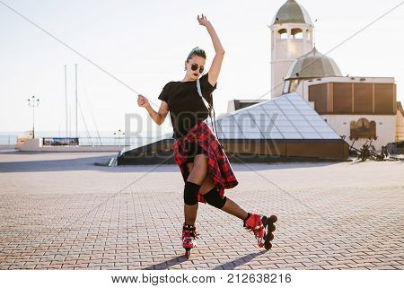 Young stylish funky girl with green hair riding roller skates and dancing near sea port during sunse
