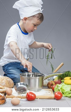 Cuisine Concepts. Portrait of Fascinated Caucasian Little Boy Working With Dill In Cooking Hat. Surrounded by Fresh Fruits and Vegetables In Studio. Posing with Smiling Expression. Against Gray. Vertical Image Composition
