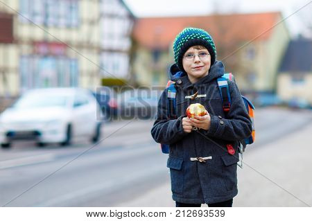Little kid boy with eye glasses on the way to the school and eating fresh apple. Child on a street. Preschooler wearing warm clothes, glasses and backpack. Happy student or people. Education concept