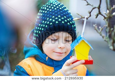 Little kid boy feeding birds in winter. Child hanging colorful selfmade bird house on tree on frosty cold day. Happy preschooler in colorful wam clothes. Selective focus on child and feeder with seeds