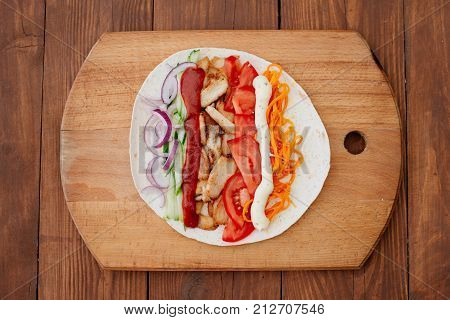 The process of preparing chiken shawarma. Ingredients sliced lie on the kitchen board. Fast food. Eastern food. Dishes of oriental cuisine lying on pita bread and decorated with greens and vegetables