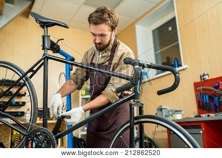 Modern repairman in uniform and earphones fixing details of bicycle