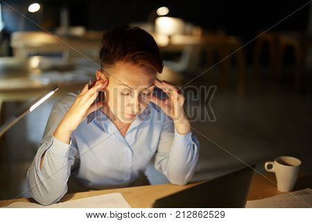 Tense businesswoman sitting in front of laptop late in the evening and preparing for report of the following day