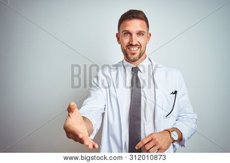 Young handsome doctor man wearing white profressional coat over isolated background smiling friendly offering handshake as greeting and welcoming. Successful business.