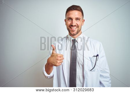 Young handsome doctor man wearing white profressional coat over isolated background doing happy thumbs up gesture with hand. Approving expression looking at the camera showing success.
