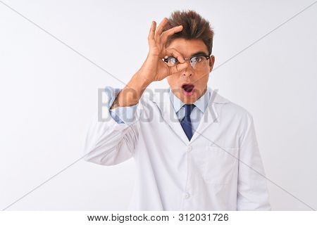 Young handsome sciencist man wearing glasses and coat over isolated white background doing ok gesture shocked with surprised face, eye looking through fingers. Unbelieving expression.