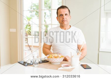 Middle age man eating asian food with chopsticks at home with serious expression on face. Simple and natural looking at the camera.