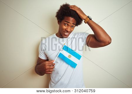 Afro american tourist man holding Argentinian Argentina flag over isolated white background stressed with hand on head, shocked with shame and surprise face, angry and frustrated. Fear and upset 