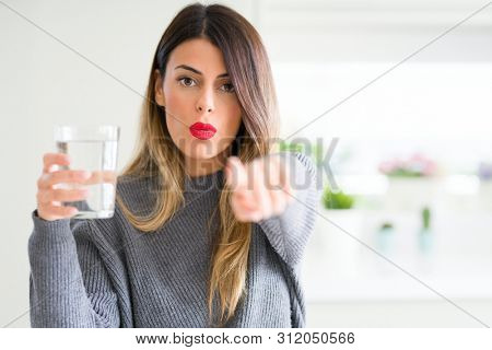 Young beautiful woman drinking glass of water at home pointing with finger to the camera and to you, hand sign, positive and confident gesture from the front